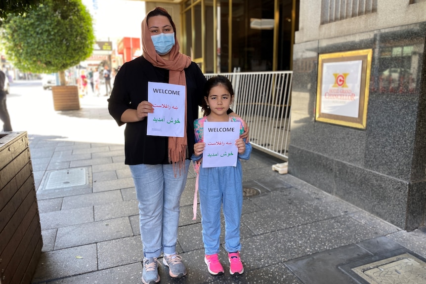 A woman and girl stand side by side holding up a sign that says 'welcome' in three languages.