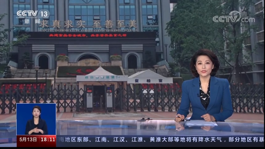 在电视银幕上，一名亚裔女主播坐在主播台上，背景投射的是一所学校的正门图片。