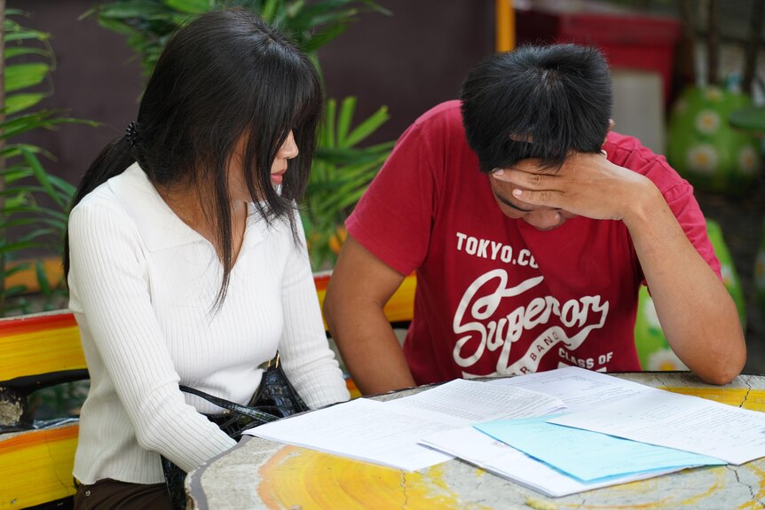 A woman in a white long-sleeved top and man in a red T-shirt sit a a table, both shielding their faces from view