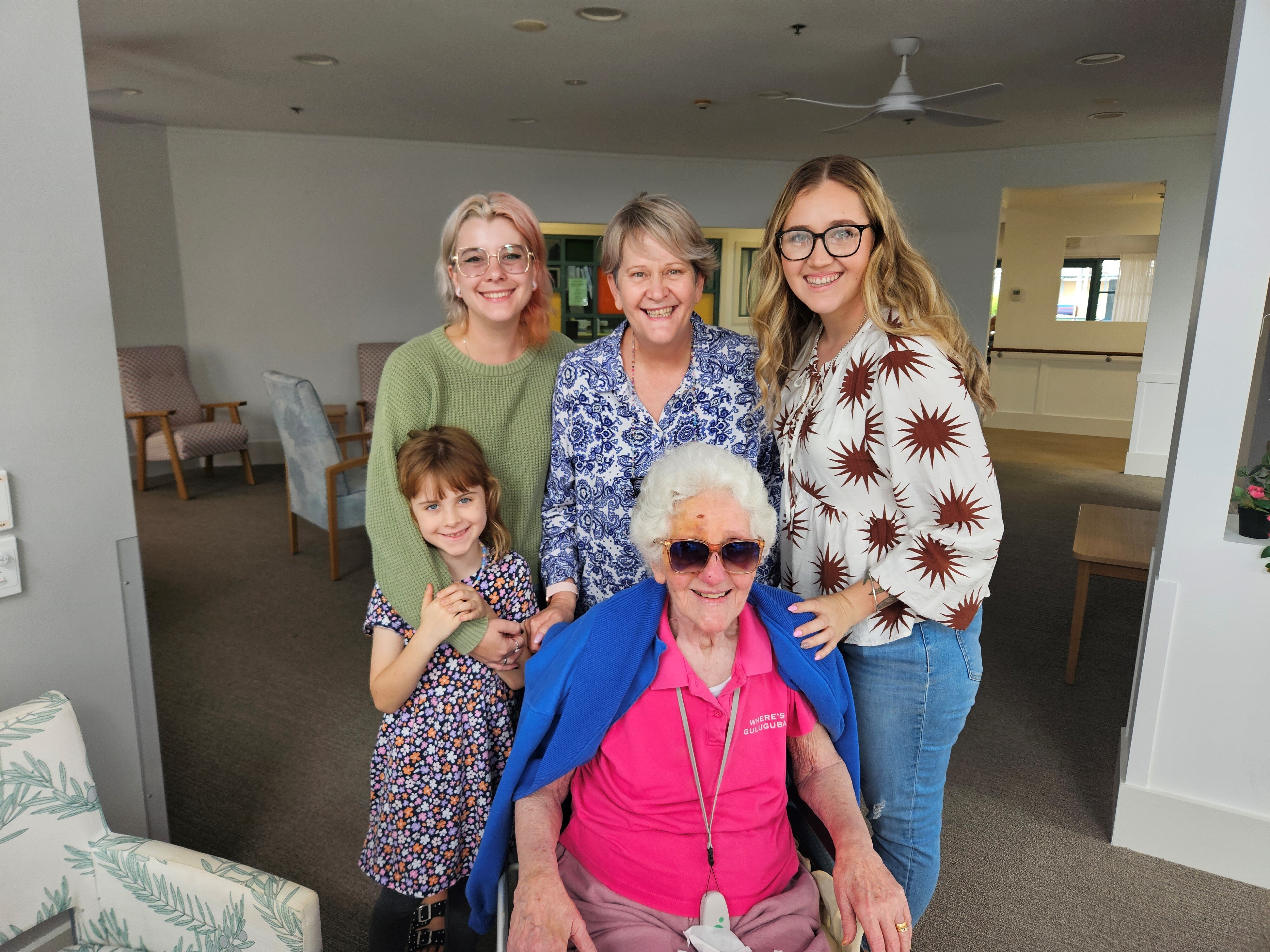 Three women and a young girl stand smiling with an older lady who is sitting.