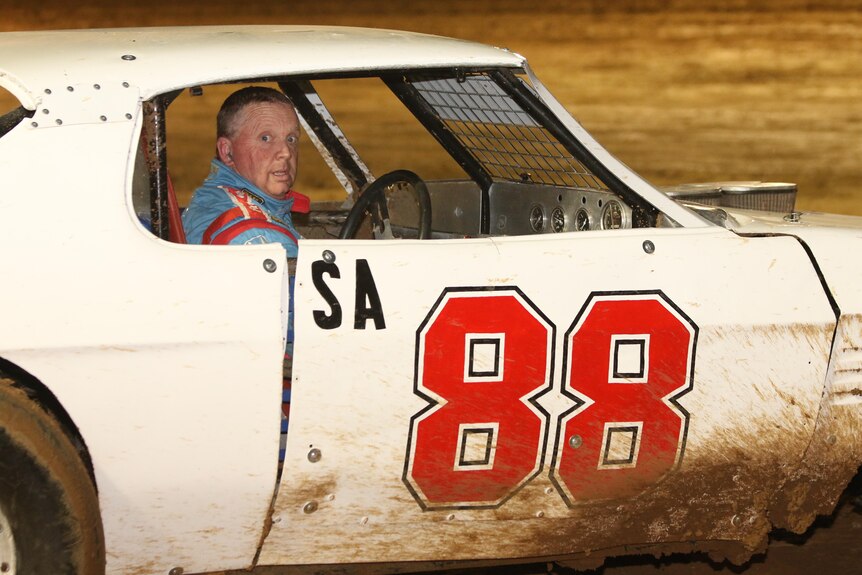 a man in blue sitting in an old white race car, a red 88 on the side of the door. Splattered mud on the underside of the car