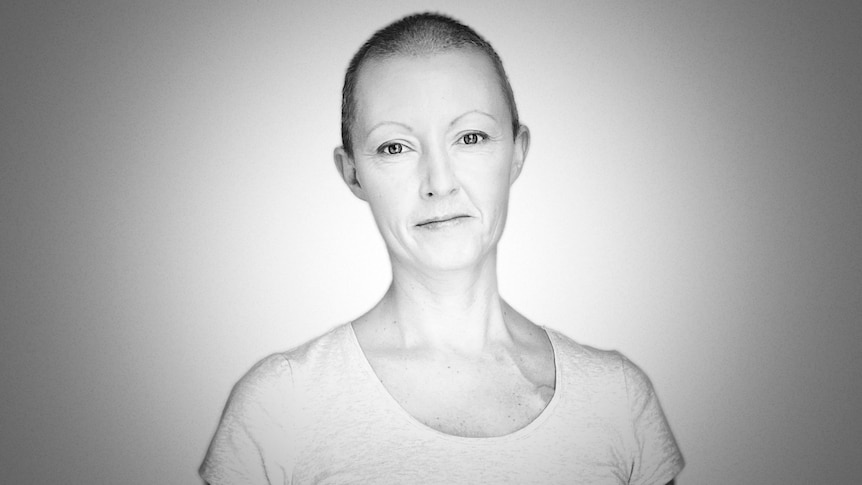 A black and white image of a woman with closely shaved hair standing against a wall.