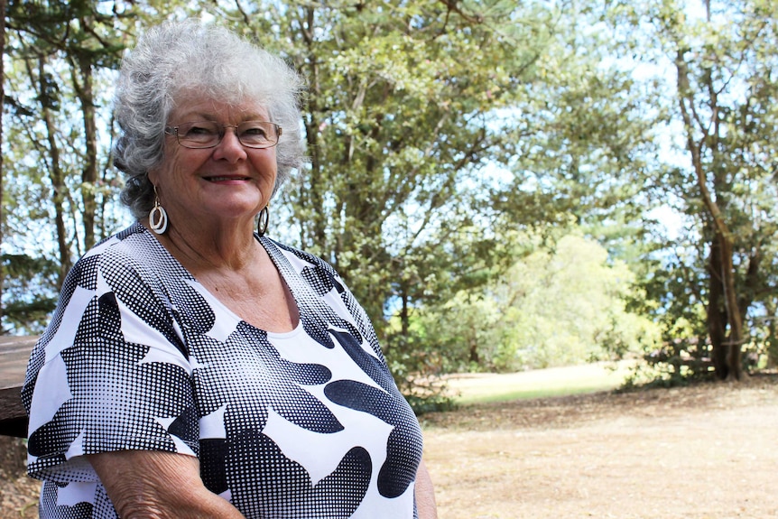 An older woman with curly grey hair stands in a forest clearing, smiling.