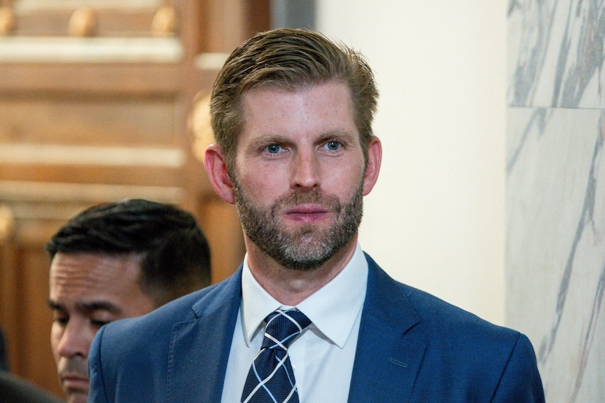 Eric Trump, dressed in suit and tie, looks straight ahead. He is sporting a beard and has product in his hair.