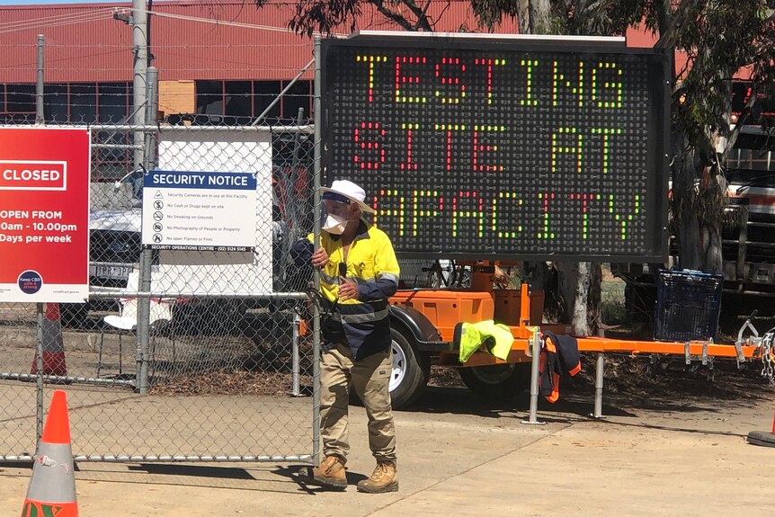 A man in hi-vis closes a gate in front of a sign reading 'Testing site at capacity'.