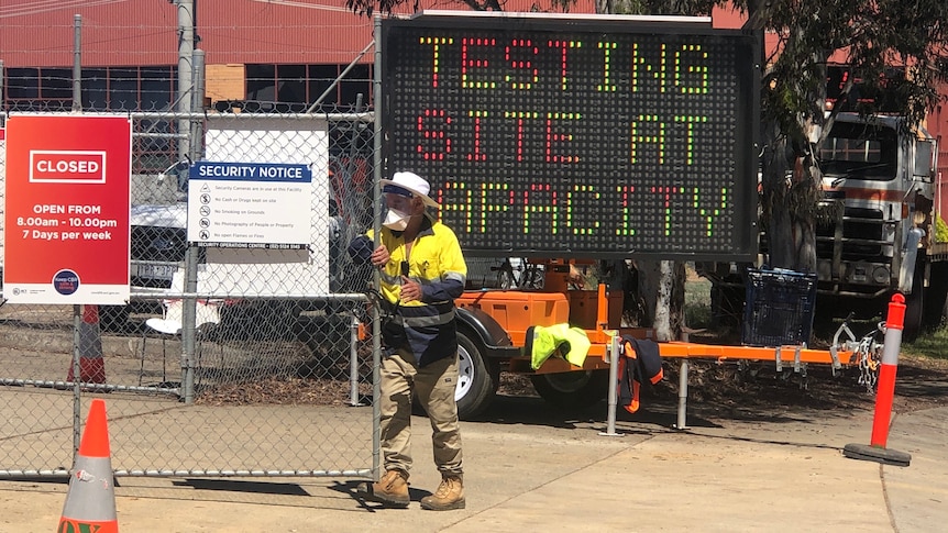 A man in hi-vis closes a gate in front of a sign reading 'Testing site at capacity'.