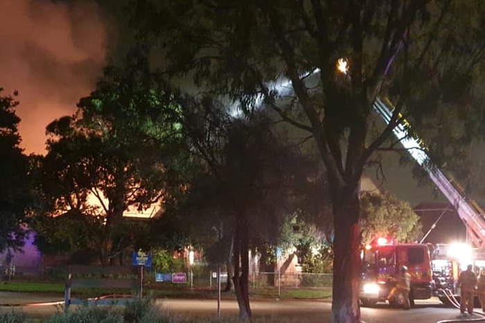 Fire lights up a dark sky and firefighters use a crane to shoot water at the blaze that's hidden behind trees.