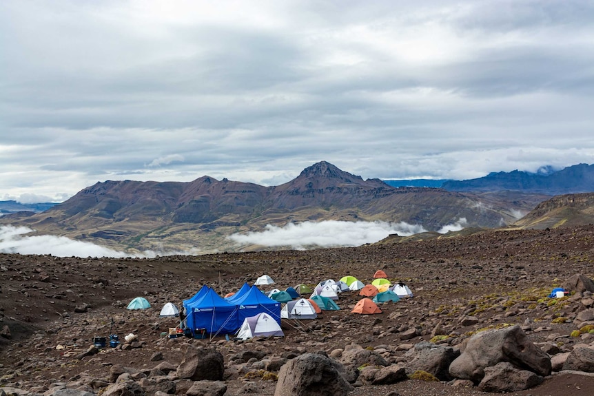 Tents sit at a rocky dig site, with mist, clouds and mountains in the background