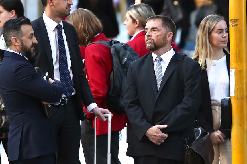 A bearded man is standing at a pedestrian crossing frowning and clasping his wrist. He wears a suit and is flanked by lawyers.