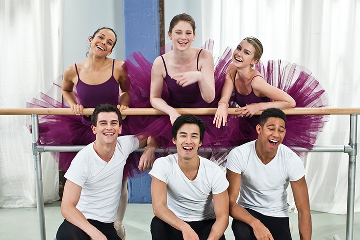 six dancers, three women wearing purple tutus and three men wearing white t shirts, laughing and smiling at the camera