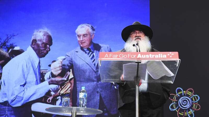 A man with a long grey beard and a hat stands at a podium with a photo of Gough Whitlam and Vincent Lingiari behind him