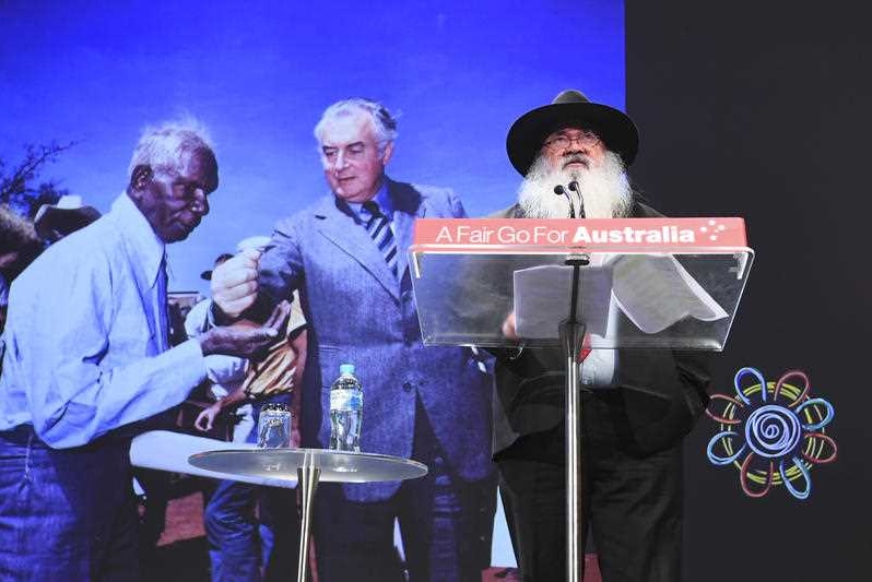 A man with a long grey beard and a hat stands at a podium with a photo of Gough Whitlam and Vincent Lingiari behind him