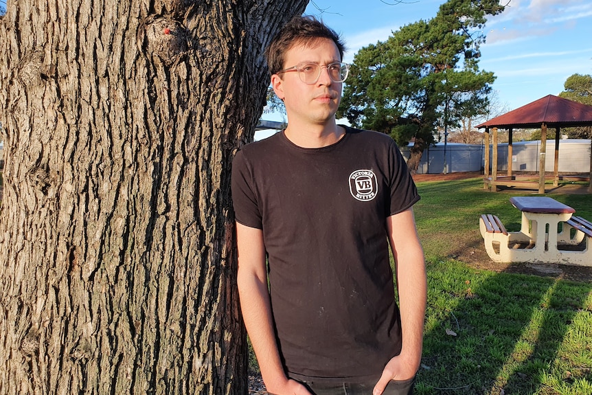 A man wearing glasses leans against a tree in a grassy area.