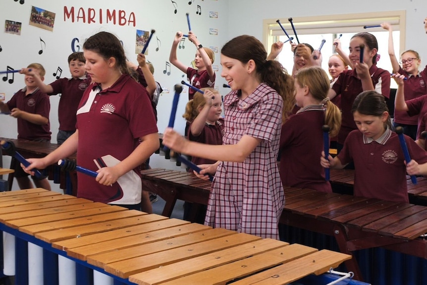 Around 20 students in maroon school uniforms play large wooden xylophones with blue sticks.