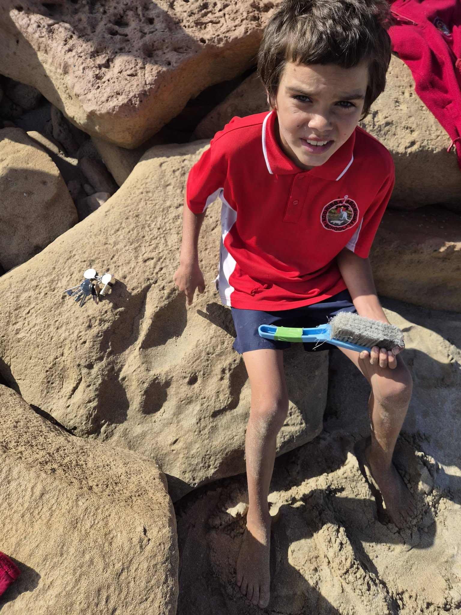 Reece, an Aboriginal boy in a red T-shirt, sitting on a rock, holding a dustpan brush. Footprint fossil in rock.