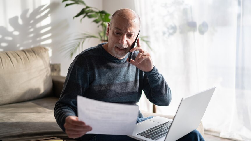 An older man talks on the phone while sitting in front of a laptop. He hold a document that he is looking at. 