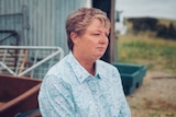 A middle-aged woman with short hair stands on a rural property and looks to the side of frame with a serious facial expression. 