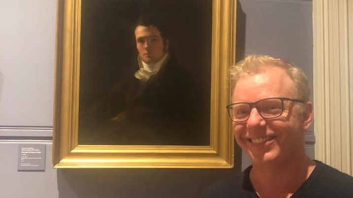 blond man with glasses stands in front of an ornately framed painting in a museum