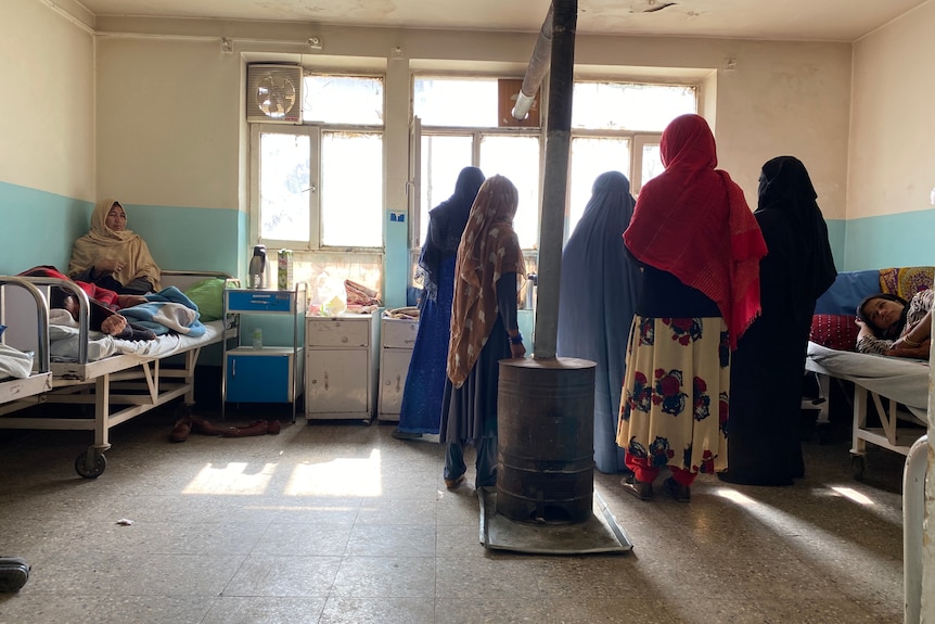 A group of women in colourful headscarves stand near a wood burner in a hospital room