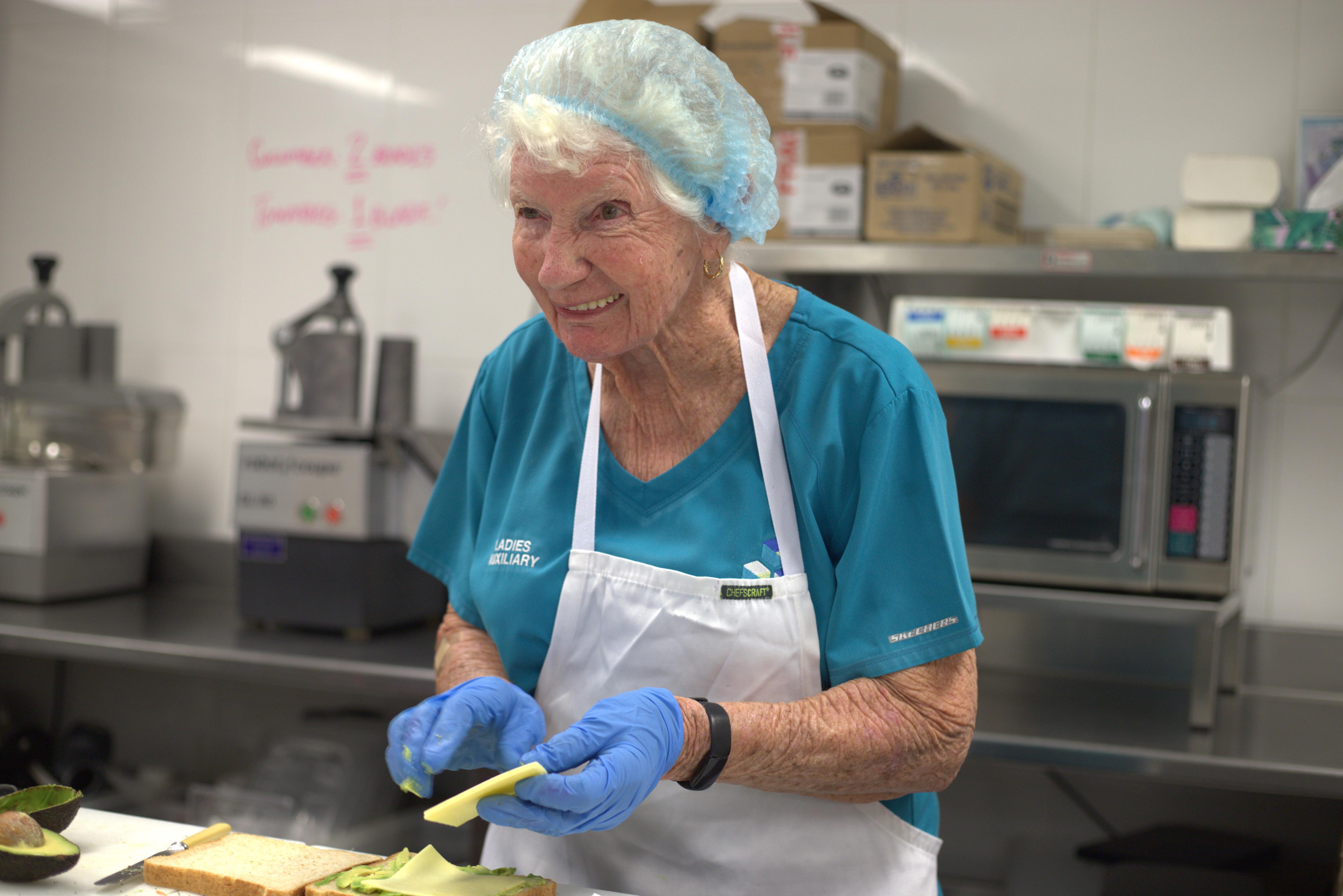 Flo is dressed in a hair net and chef clothes, preparing a sandwich, smiling