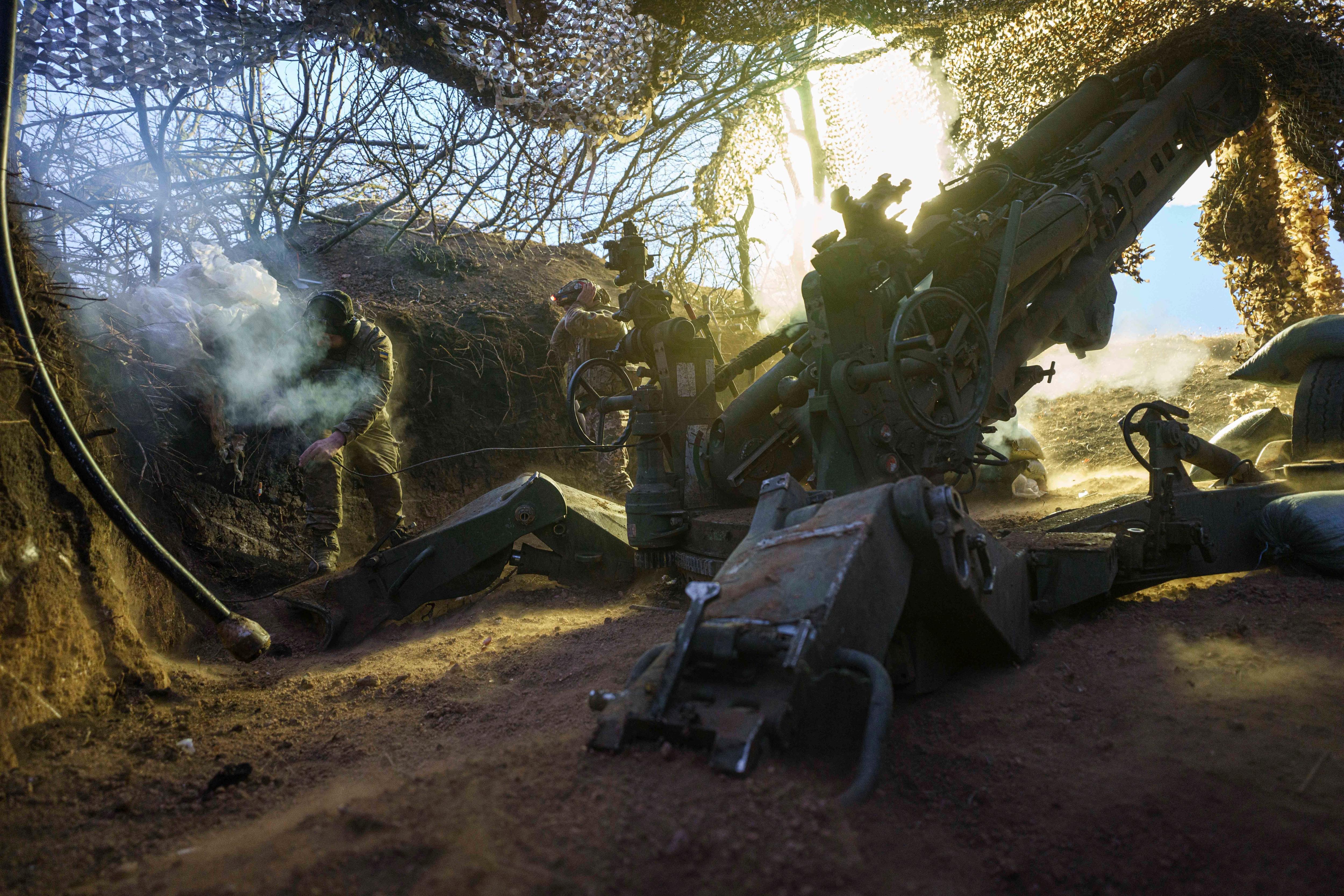 Soldiers lit by early morning light, with fog, smoke and a camouflage net overhead.