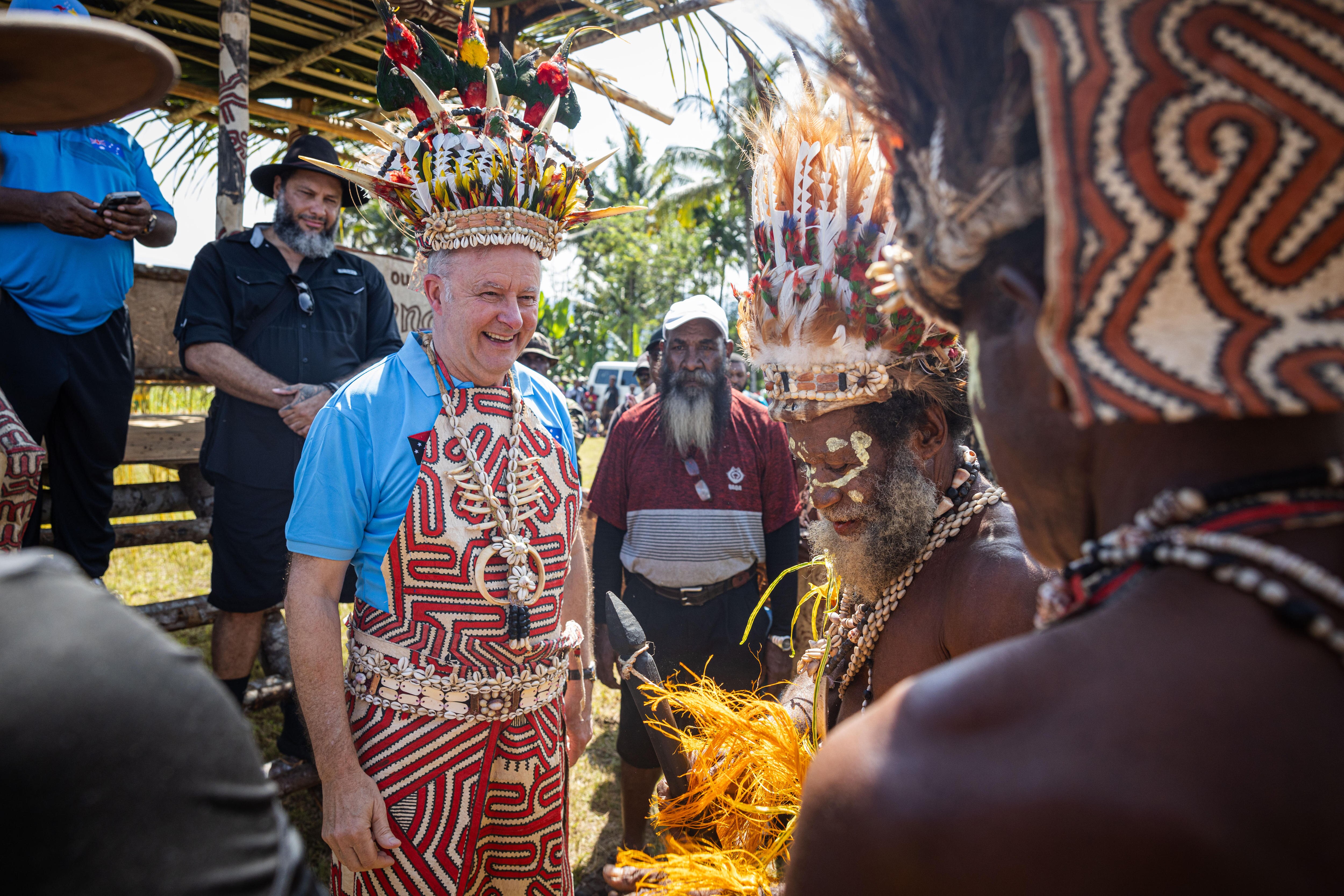 Albanese wears a feathered headdress and patterned clothing, smiling, surrounded by Papuan men.