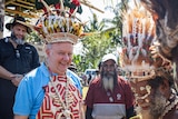 Albanese wears a feathered headdress and patterned clothing, smiling, surrounded by Papuan men.