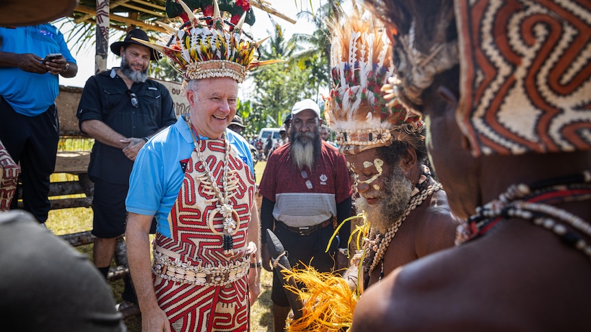Albanese wears a feathered headdress and patterned clothing, smiling, surrounded by Papuan men.