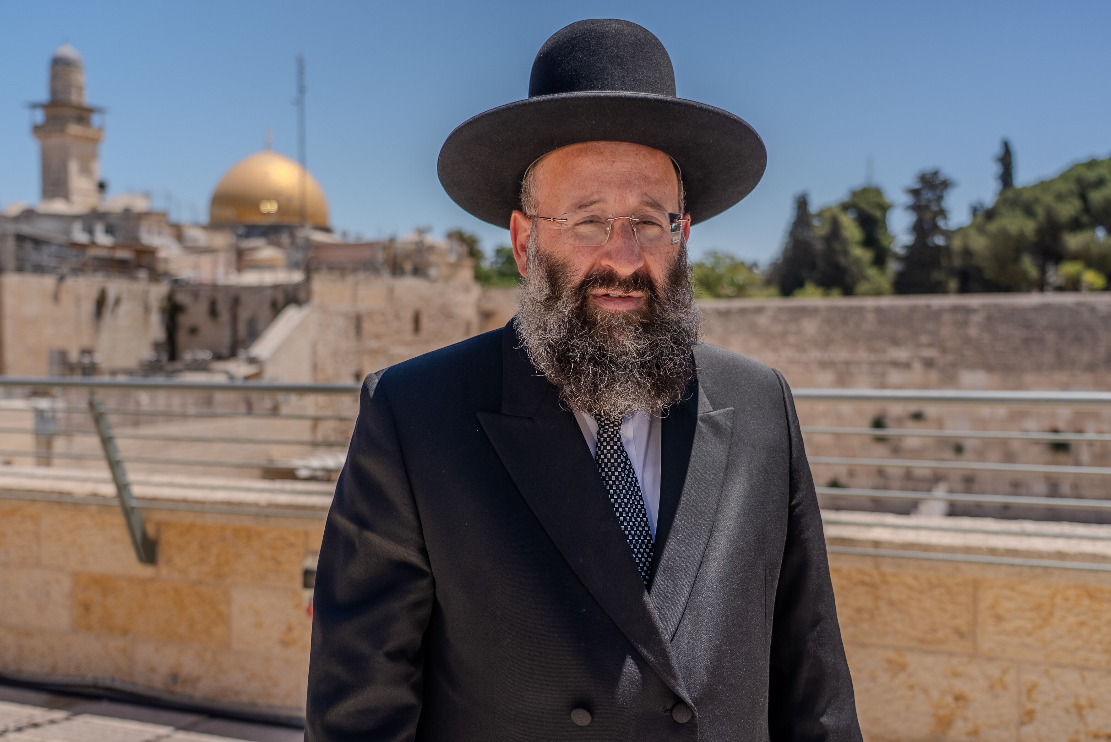 Rabbi Shmuel Rabinovich wears a black hat and black suit as he stands in bright sunlight with a large stone compound behind.