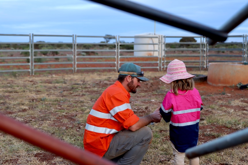 Un hombre y su hija pequeña con lentes de alta visibilidad mirando un teléfono dentro de un corral de ganado.