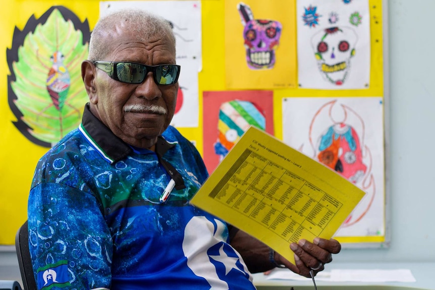 A teacher wearing sunglasses holding up a book in a classroom with drawings in the background.