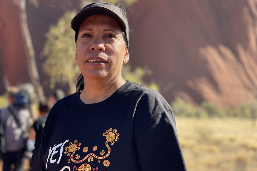 A woman stands at the base of Uluru, wearing a cap and T-shirt. 