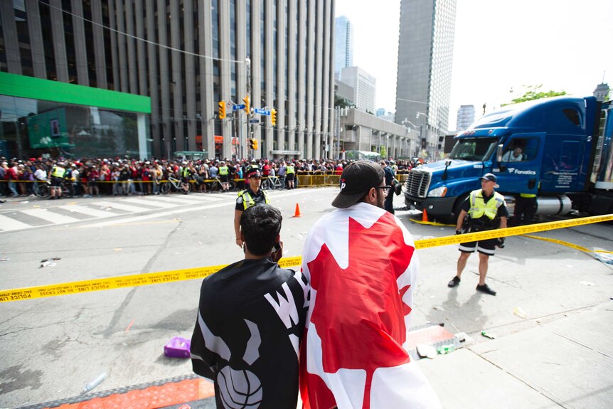 A man with a Canadian flag wrapped around his shoulders stands by crime scene tape, watching police officers.