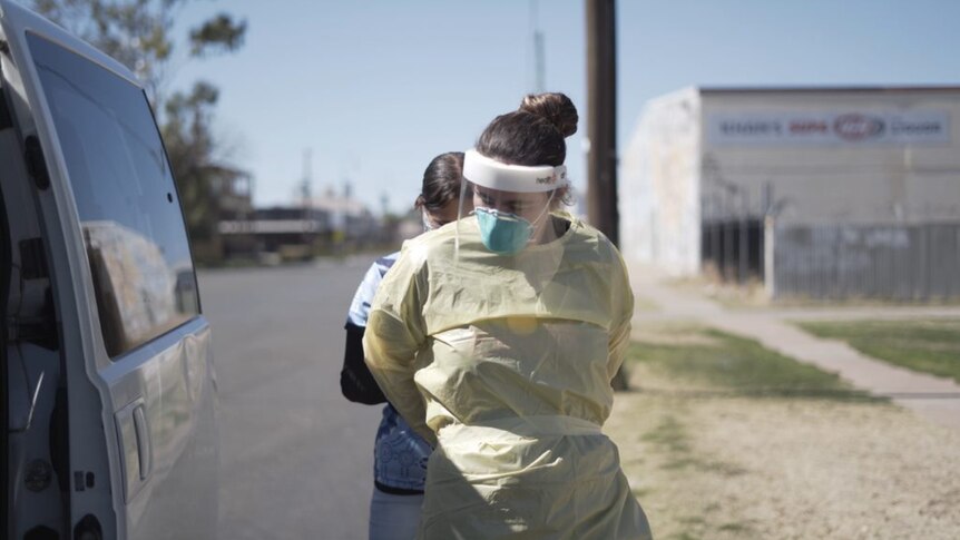A female health worker helps another tie up her PPE next to a parked van.