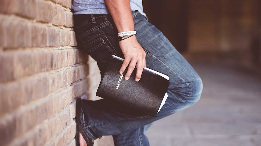 The lower half of a young man's body leans against a wall while holding a bible.