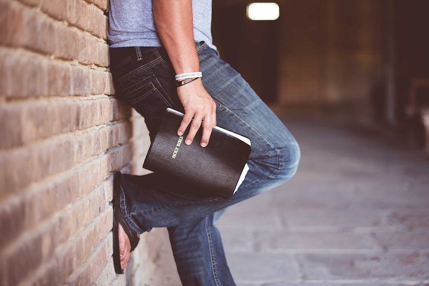 A stock photo shows a young man leaning against a brick wall and holding a Bible.