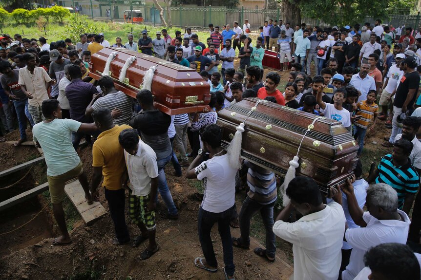 A crowd of people stand as men carry two wooden coffins.