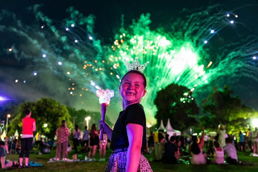 A young girl holds a wand and smiles as New Year's Eve fireworks erupt behind her