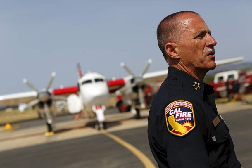 A fireman with a close-shaved head stands on the tarmac in front of an aircraft on a sunny day.