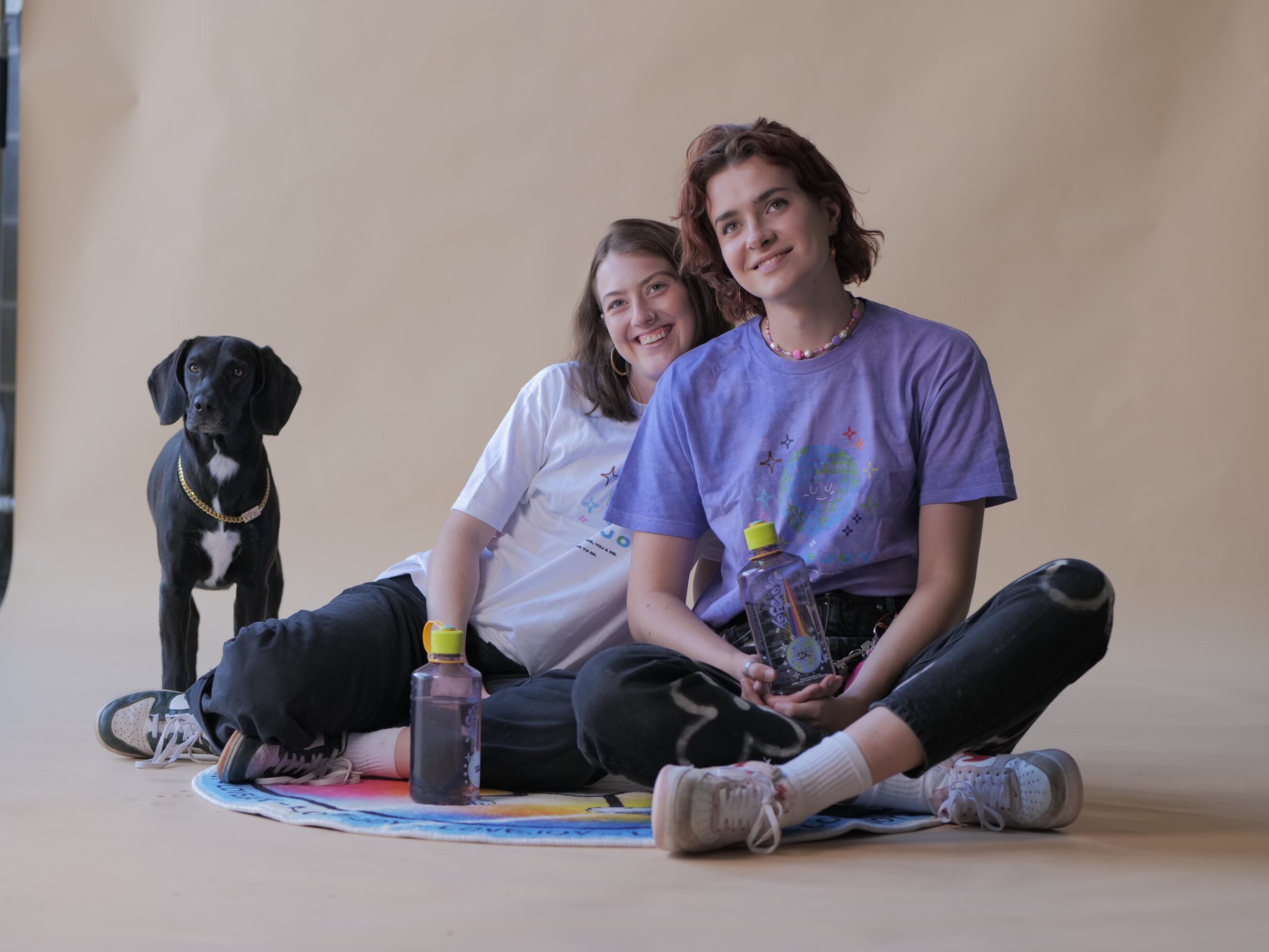 Queenie's black dog is seen left, with Queenie sitting in the middle and Jan on the right. They smile and look left.