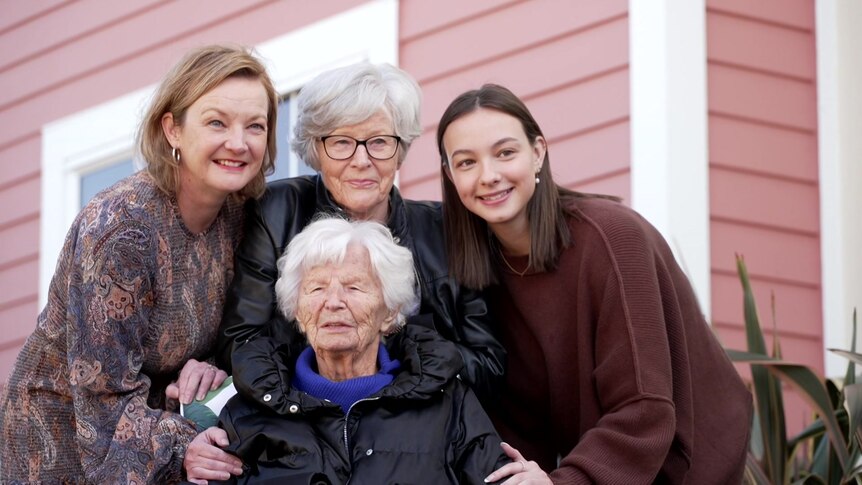 Three women are standing around a white-haired woman who is seated