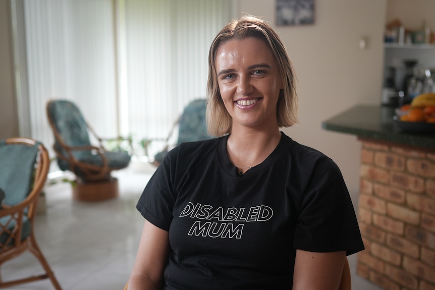 A young white woman with short blonde hair smiling. She is sitting in a lounge room and wearing a shirt that says 'disabled mum'