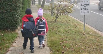 Two primary school aged children wearing schoool uniforms in winter walk along a sidewalk