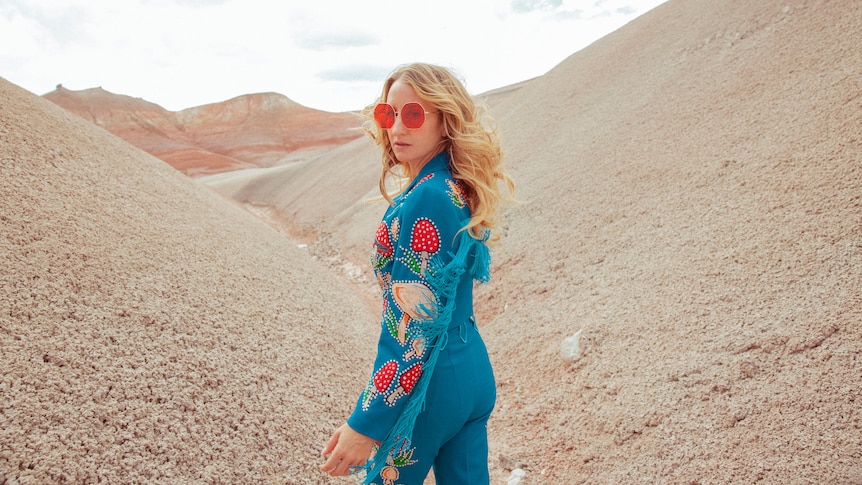 Margo Price stands in a dessert with red sunglasses on and a blue jumpsuit