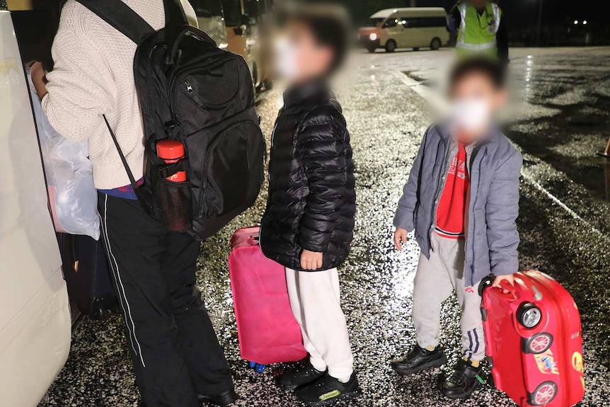 Adults and children with luggage line up to board a bus at night.