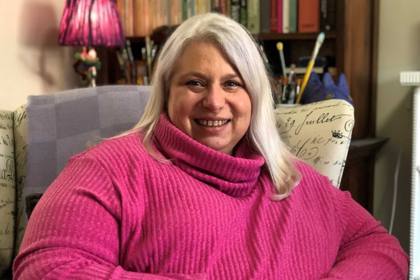 A woman with grey hair wearing a pink top sits in a chair.