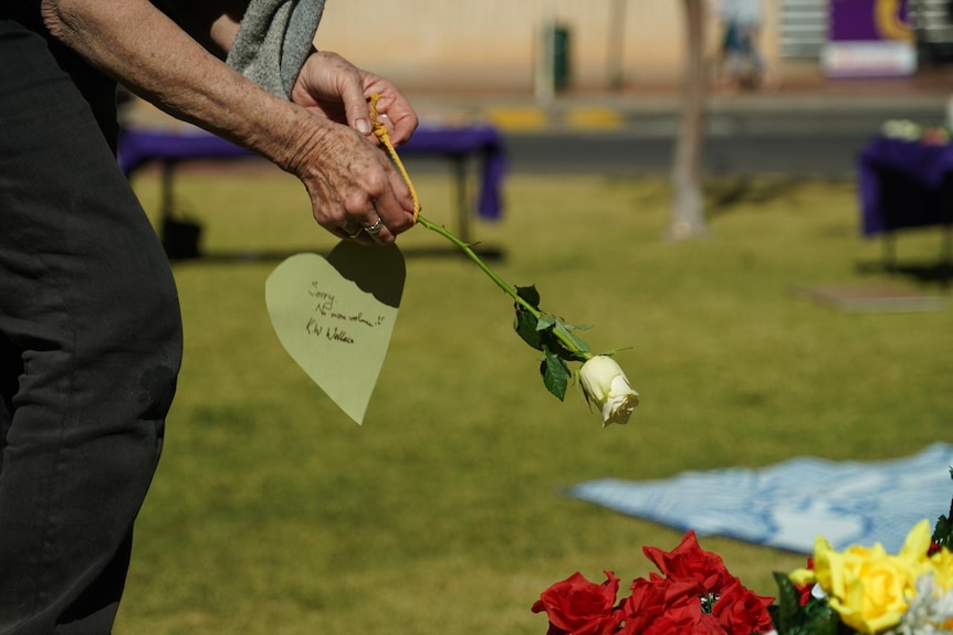 A woman's hand holds a white rose with a note attached to it which reads 'sorry, no more violence.' 