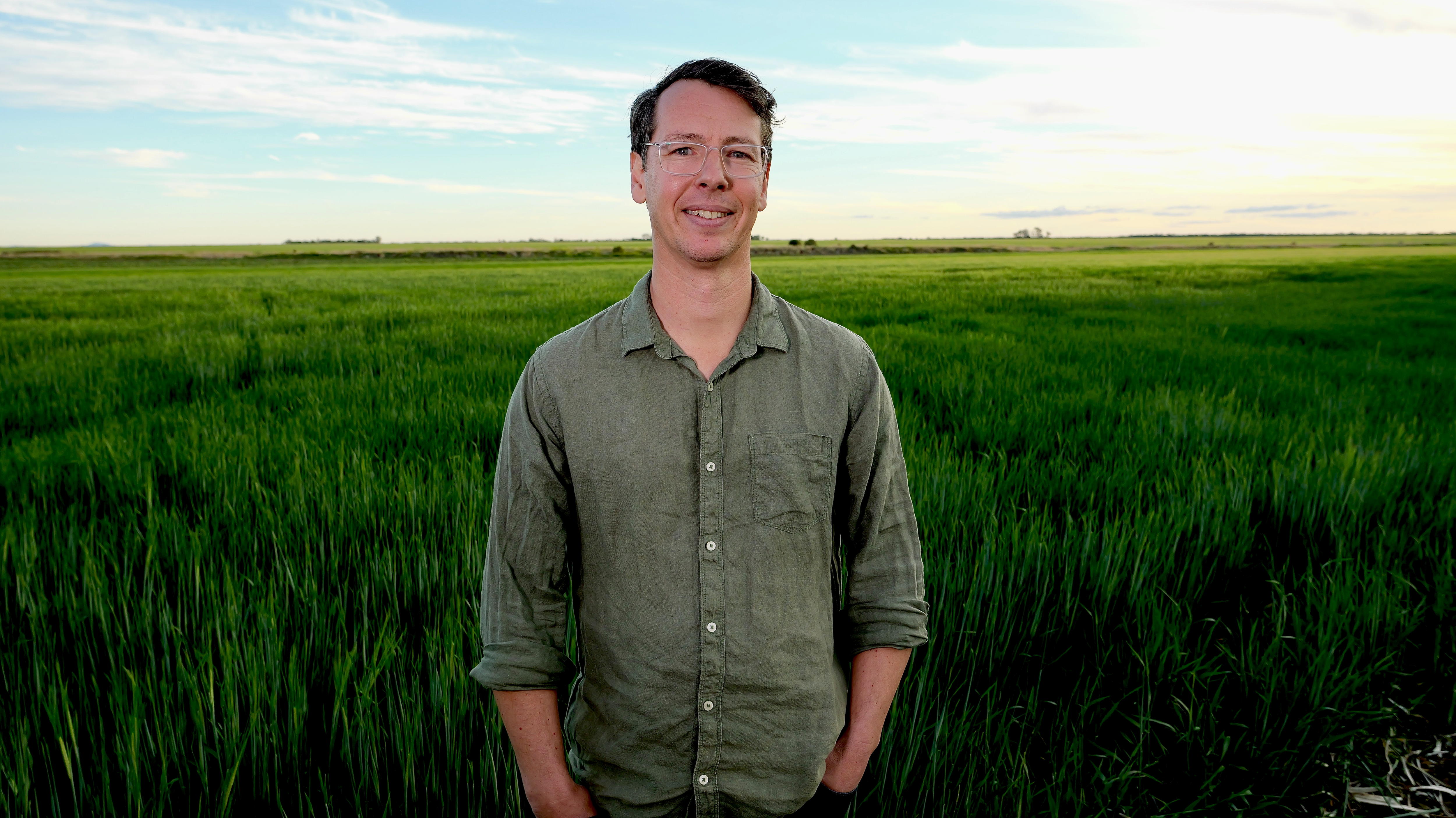 A man with a button up, green shirt stands smiling in front of a green crop.