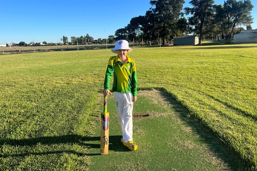 A young boy holding a cricket bat wearing cricket gear and standing on a cricket pitch