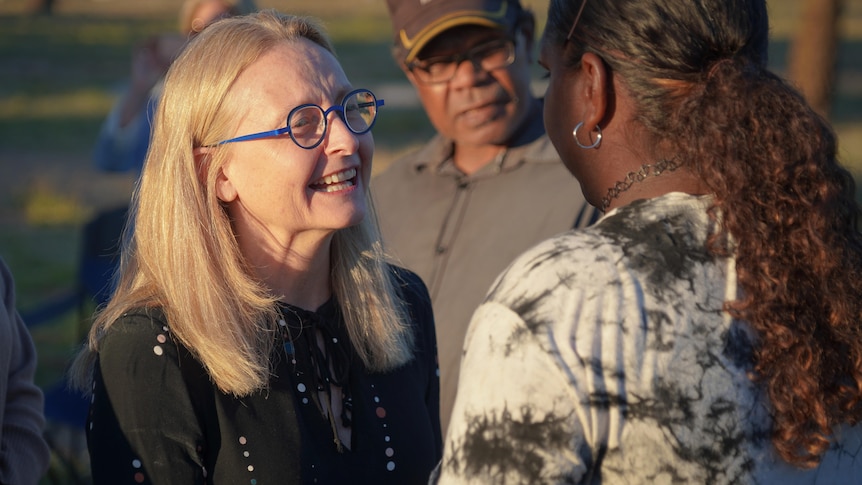 A white woman with blonde hair and glasses smiles as she speaks to an indigenous woman 